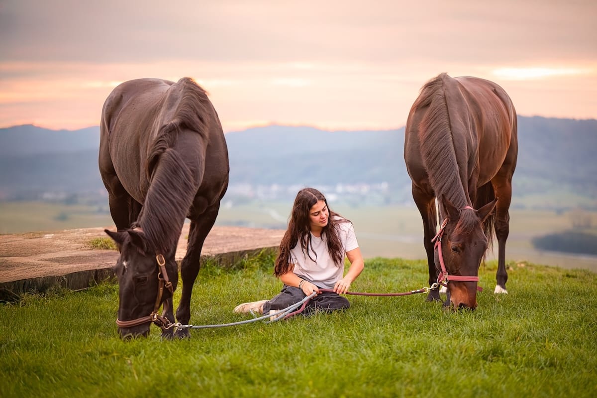 Jeune femme assise dans l'herbe avec ses deux chevaux qui broutent paisiblement